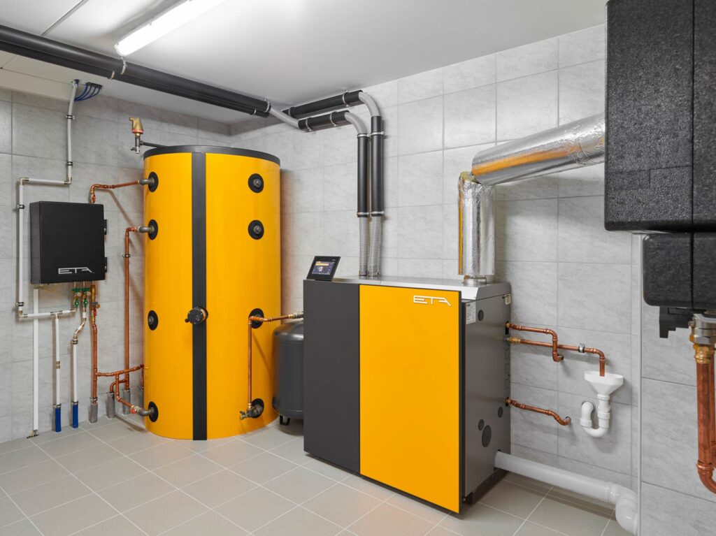 Ein moderner Heizraum mit gelbem Boiler und Steuerung; silberne Rohre und Fliesenwände.